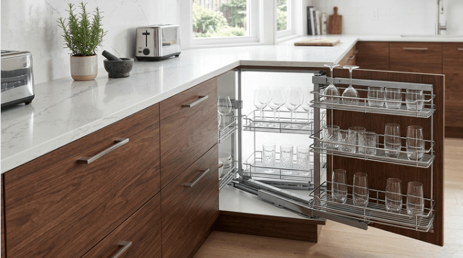 A premium multi-tier chrome magic corner storage system smoothly sliding out from an L-shaped walnut kitchen cabinet, neatly holding modern glassware.