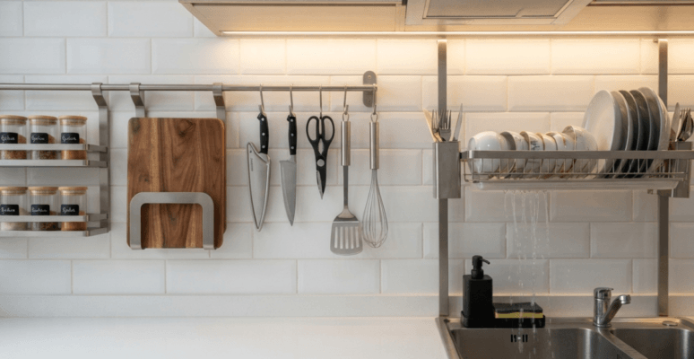 A close-up of a premium stainless steel hanging kitchen organizer rail holding spices, a cutting board, and utensils above a clear countertop in a modern BD kitchen.