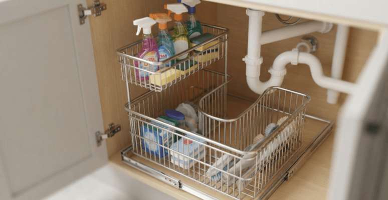 A close-up, high-angle photograph of a premium SUS304 stainless steel under sink storage basket, perfectly fitted around PVC plumbing in a modern Bangladeshi kitchen cabinet.